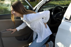 Woman holding her lower back while getting out of a car, representing muscle pain after a car accident.
