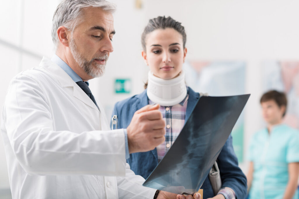 Doctor reviewing spinal X-rays with a patient wearing a neck brace after a car accident, discussing a possible vertebral compression fracture.