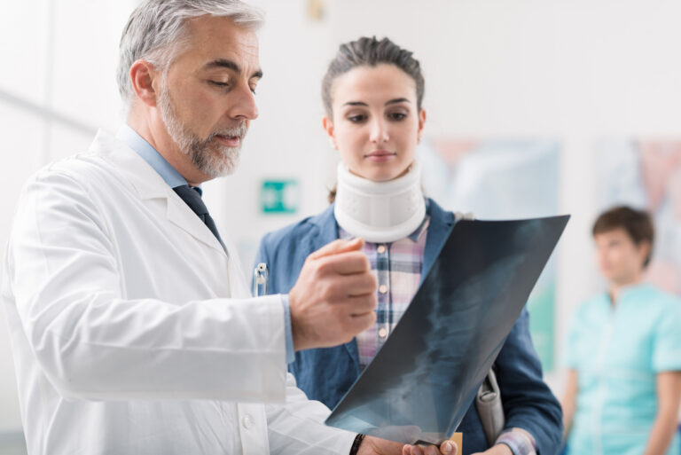 Doctor reviewing spinal X-rays with a patient wearing a neck brace after a car accident, discussing a possible vertebral compression fracture.